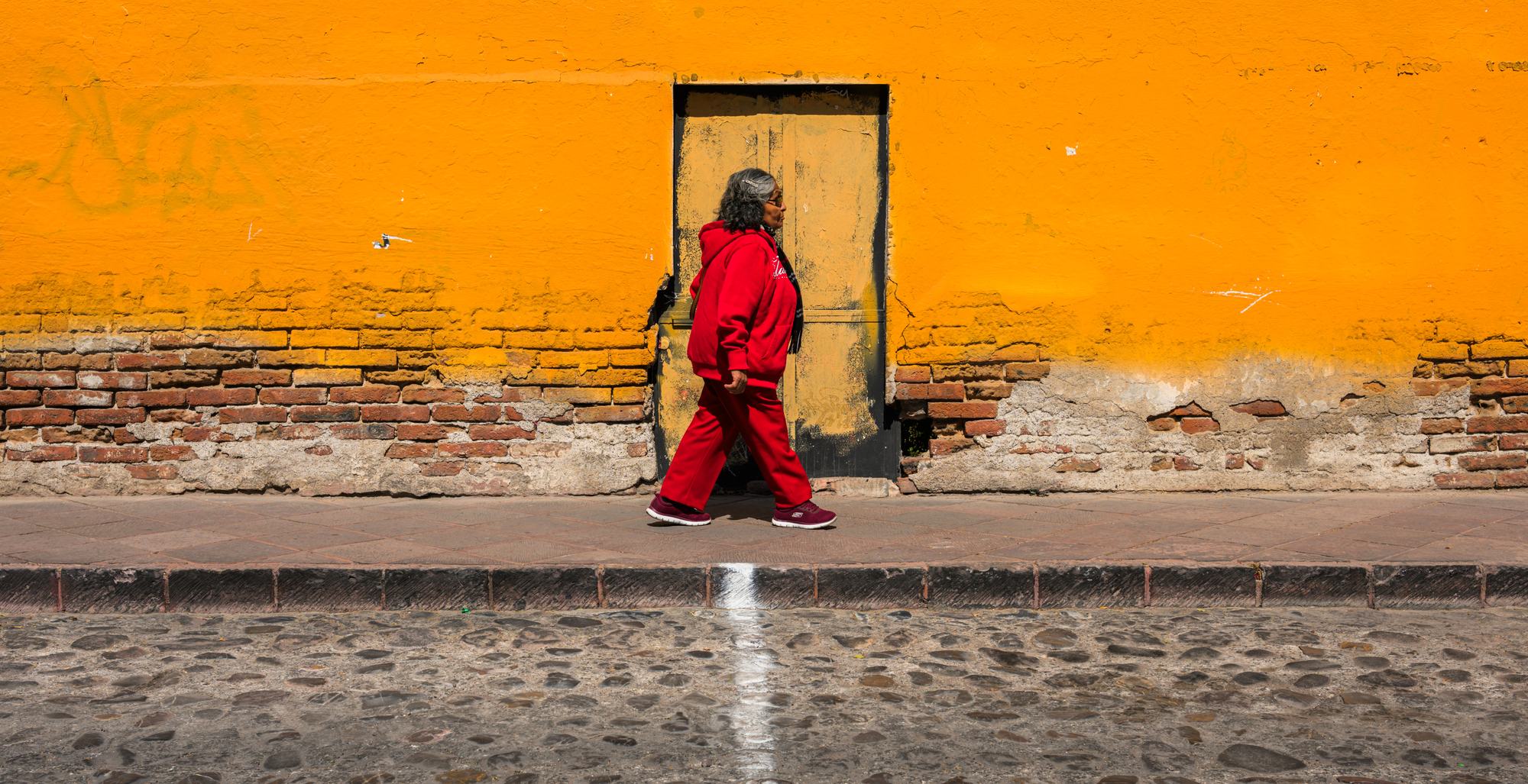 Street photography in San Miguel de Allende: a woman in red walking past an orange wall. WorthWait, 35mm