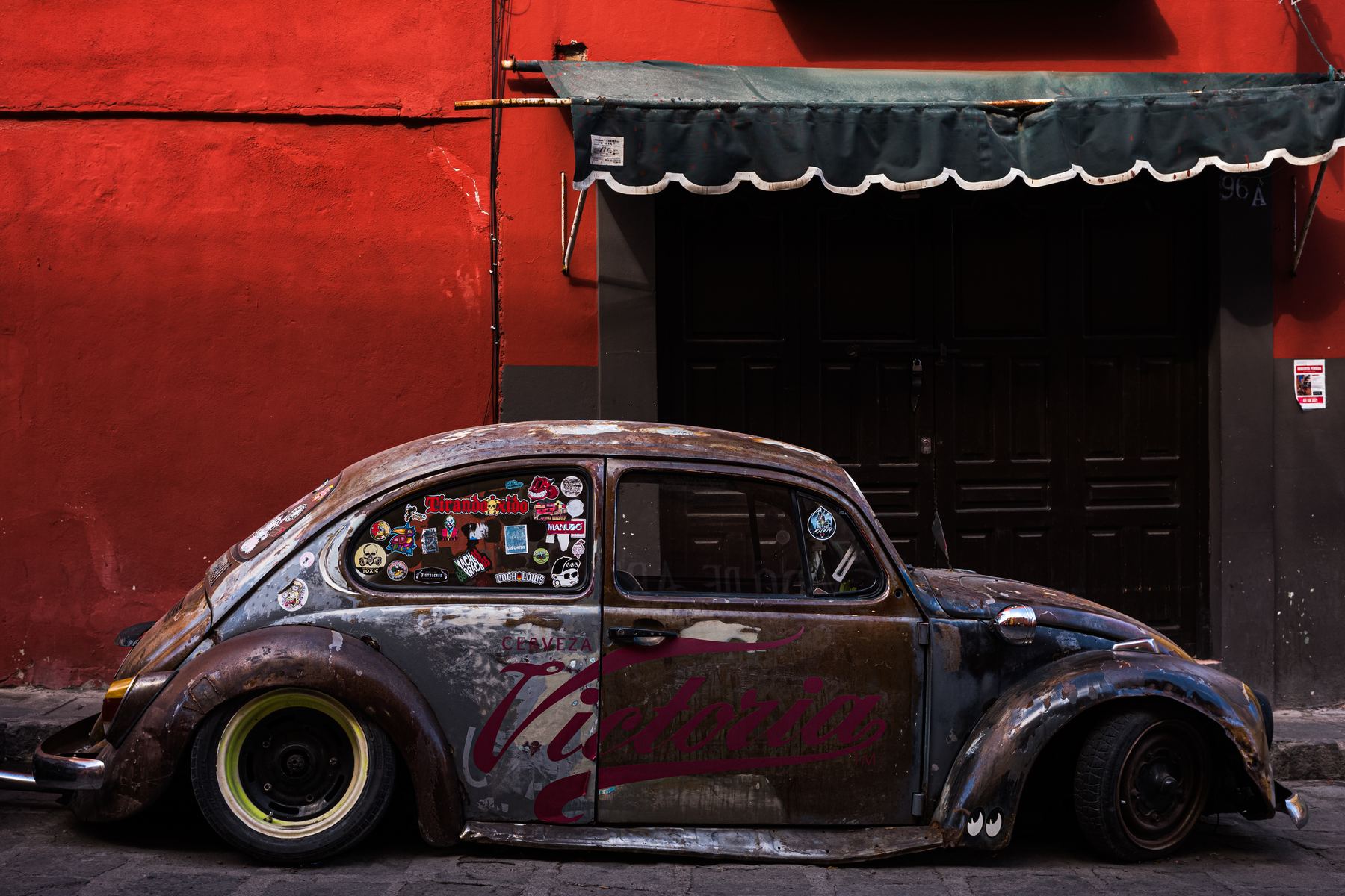 35mm street photography: a rusted Volkswagen Beetle covered in stickers, parked against a red wall