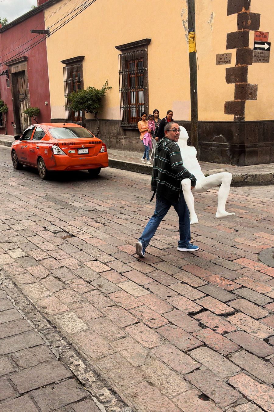 Disposable camera app for street photography: a man carries a white mannequin across a cobblestone street, Negative film