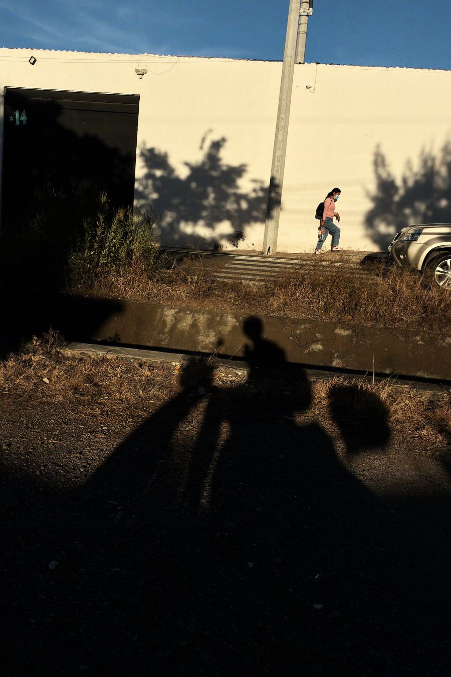 Candid street photography on iPhone: the shadow of a man on a bicycle stretches across a dark lot as a woman walks past a sunlit wall, Negative film