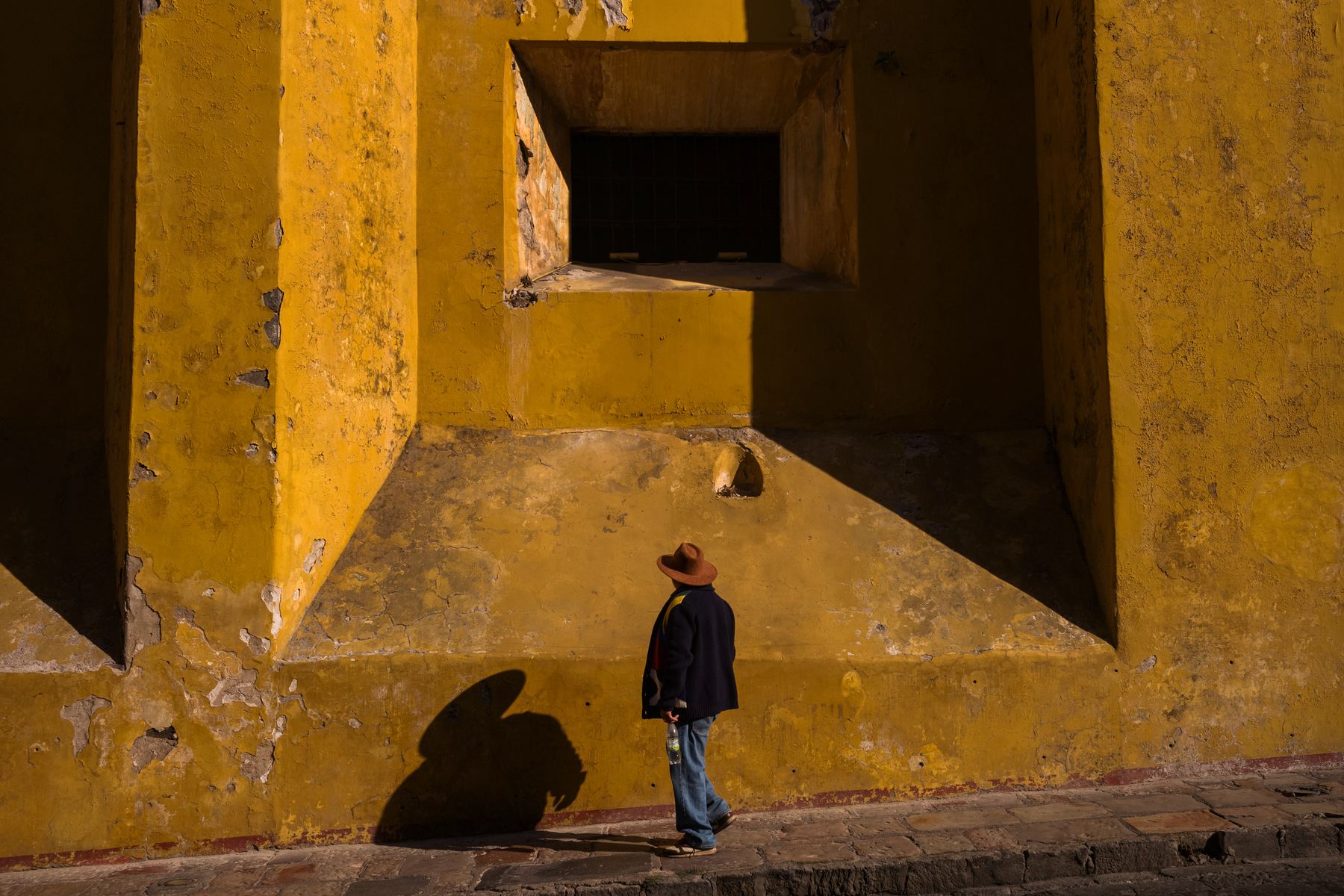 Street photography at 35mm: a man in a hat waits by a weathered yellow wall, his shadow cast on the stone